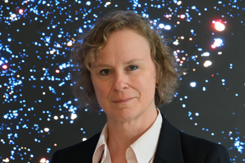 A white woman with blonde, curlyhair and wearing a white shirt and dark jacket, stands in front of a starfield
