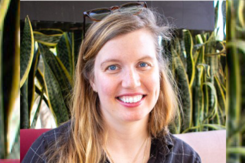 A white woman with fair hair and wearing a blue shirt sits in front of a leafy background