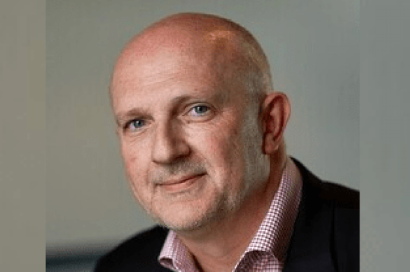A white bald man wearing a dark jacket and pink shirt, sits in front of a green background