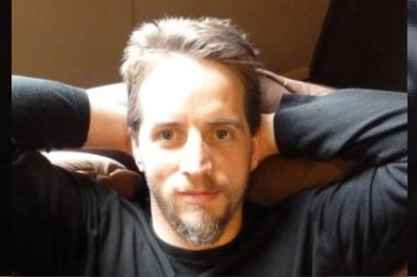 A white  man with fair hair and a beard sits with his arms behind his head