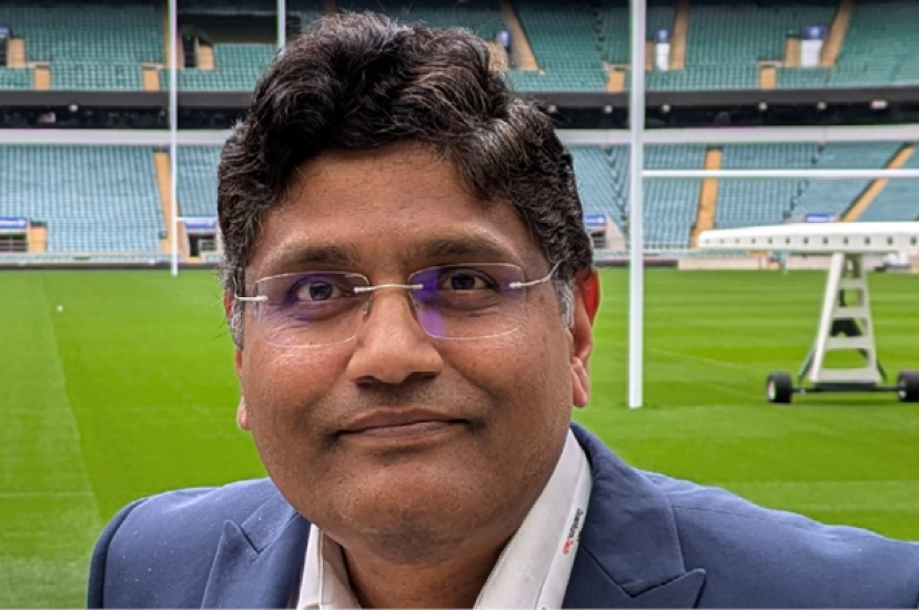 A Asian man with dark hair wearing a suit jacket sands in front of a rugby pitch