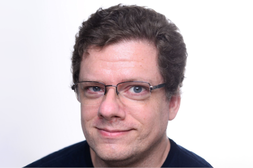 A white man with glasses and curly hair wearing a blue t-shirt
