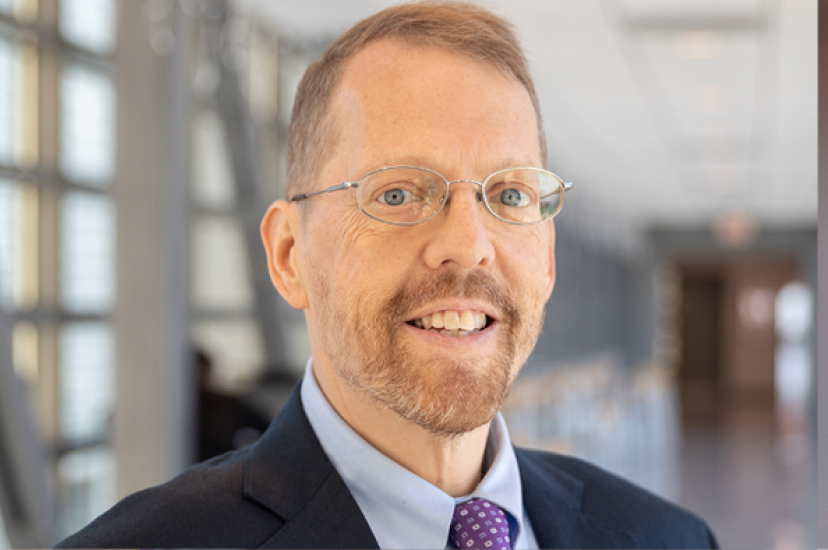 A white man with fair hair and a beard wearing a suit in a brightly-lit corridor