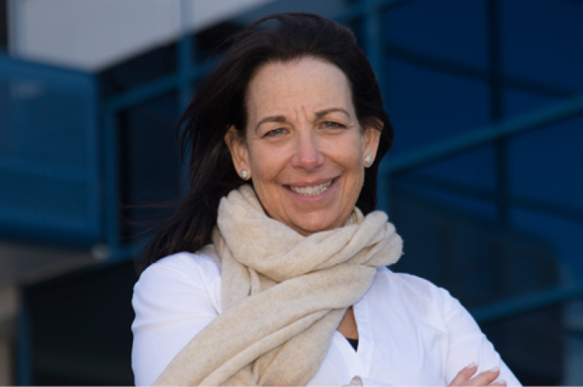 A white woman with brown hair and wearing a white shirt and cream scarf stands in front of a glass building