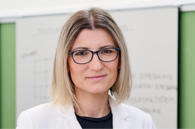 A blonde woman with glasses wearing a lab coat stands in front of a whiteboard