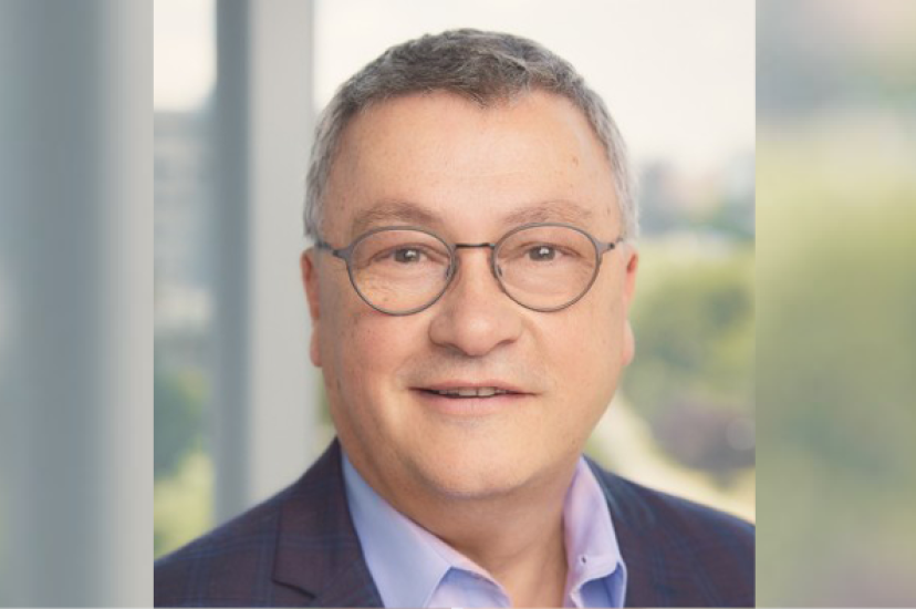 A white man with greying hair and glasses is wearing a suit and standing in front of a window