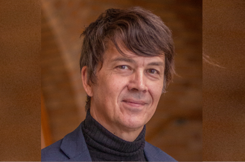 A white man with greting hair and wearing a grey jacket stands in front of a brown background