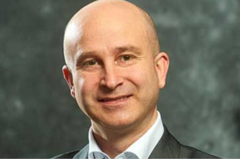 A bald white man wearing a suit stands before a green background 
