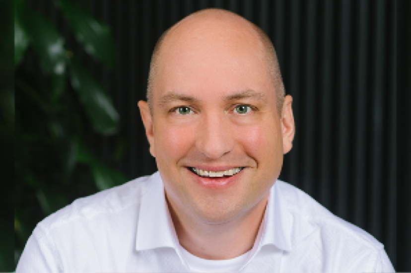 A bald white man wearing a white shirt sits before a dark background with a plant to his left