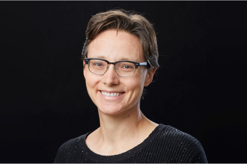 A woman with short brown hair and glasses wears a dark jumper and sits in front of a dark background