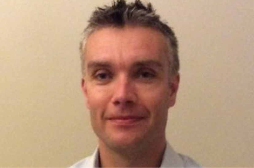 A white man with short greying hair stands in front of a beige background