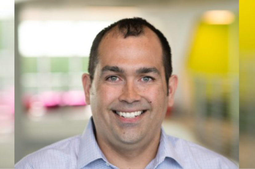 A white man smiles at the camera. He is wearing a striped shirt 