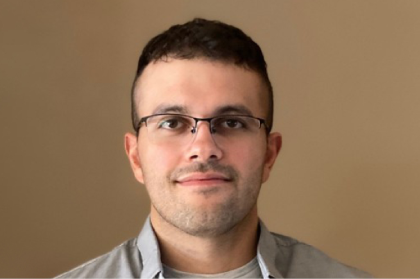A white man with short dark hair and glasses wearing a grey shirt stands before a brown background