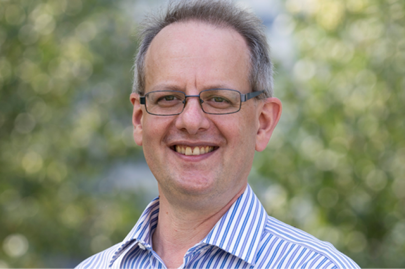 A white man with glasses and wearing a striped shirt, stands before a leafy background