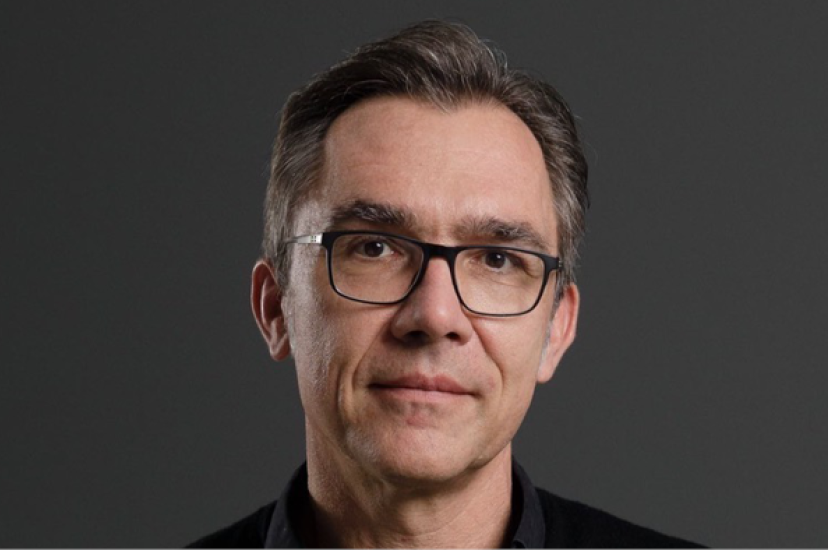 A white man with glasses and wearing a black shirt stands in front of a grey background