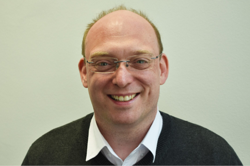 A white man with glasses and wearing a white shirt and jumper sits in fronto og a green background