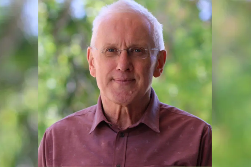A man with white hair wearing a red shirt stands in front of a leafy background