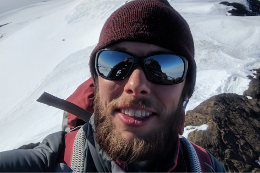 A white man witha beard wearing a hat and sunglasses takes a selfie at the top of a mountain