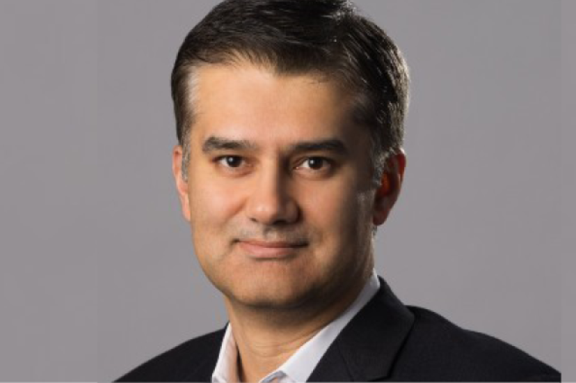 A man with dark brown hair and a dark suit stands in front of a grey background