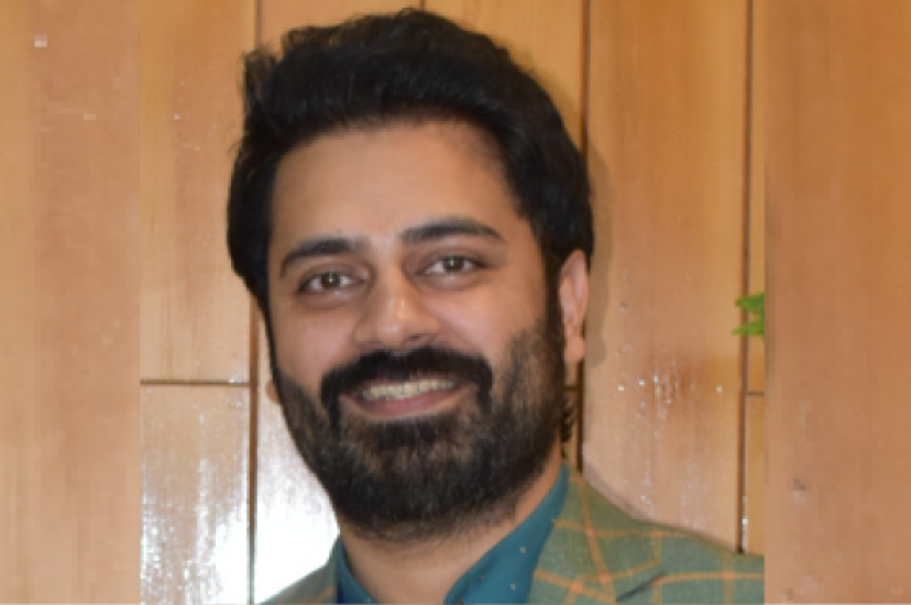 An Asian man withdark hair and a beard is wearing a green check suit and standing before a woodpanelled wall