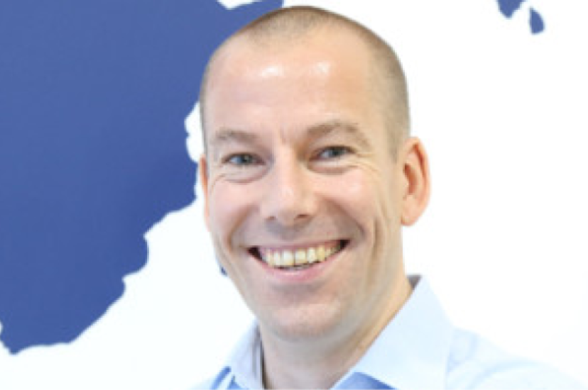 A smiling white man with a shaved head and wearing a light blue shirt stands in front of a map mural