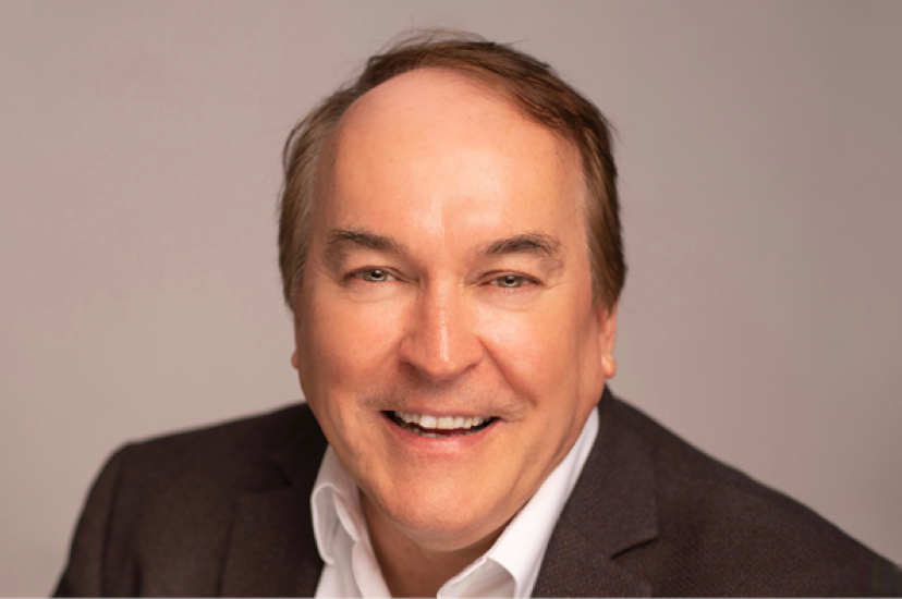A white, clean shaven man wearing a brown suit sits in front of a beige background
