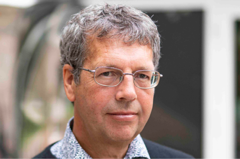 A white man with greying hair and glasses, wearing a shirt and jumper