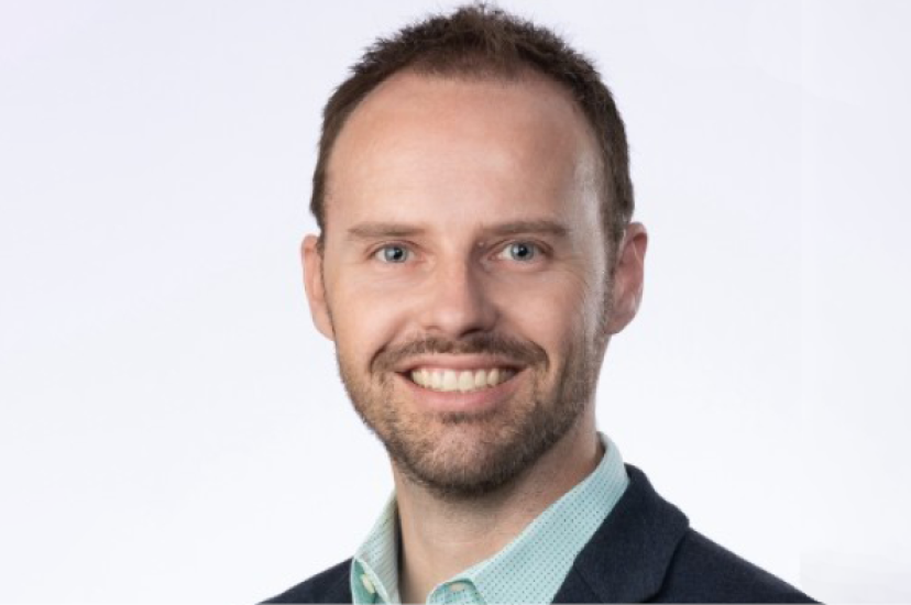 A white man with a light beard smiles at the camera. He is wearing a blue suit and a green shirt 
