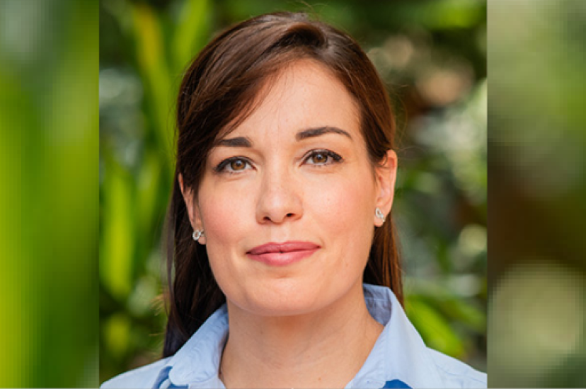 A white woman with long brown hair and brown eyes wears a blue shirt in front of some green foliage