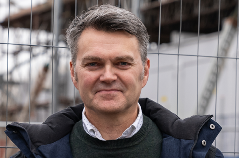 A white man with greying hair wears a blue raincoat in front of a chainlink fence