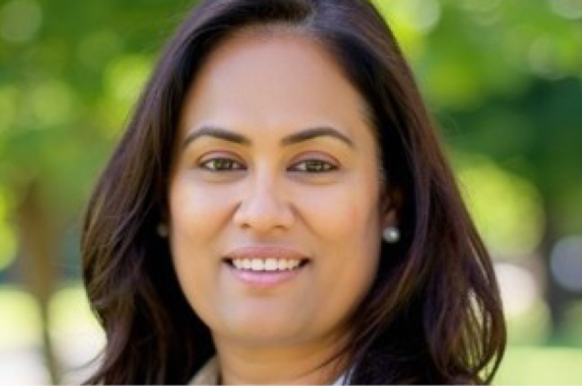 A woman with dark brown hair and brown eyes wears a white shirt. She is standing in front of a leafy park