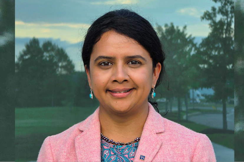 An Asian woman with dark brown hair and a pink jacket stands in front of a leafy background