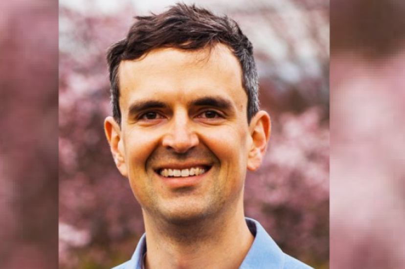 A white man with short brown hair. He is smiing and standing in front of some red trees