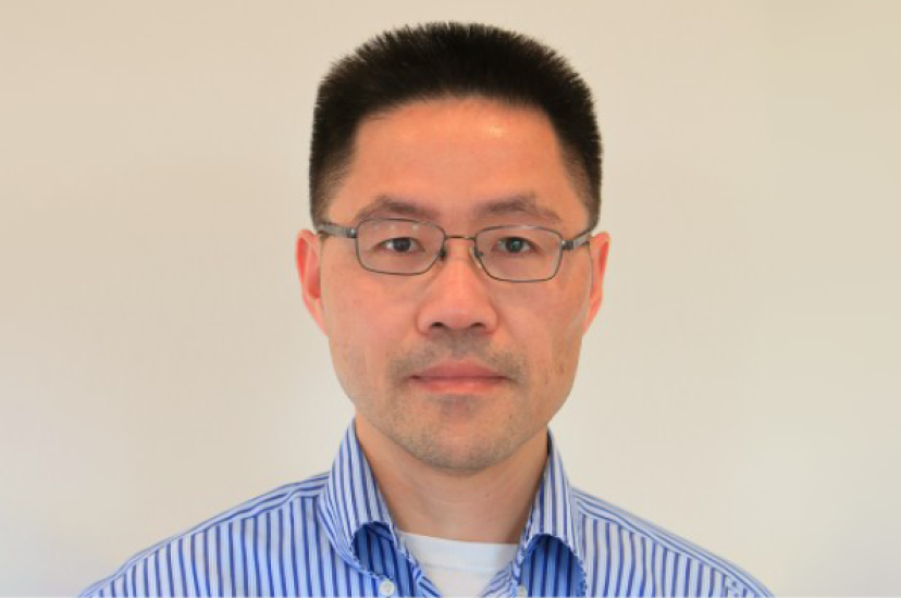 An Asian man with glasses and wearing a blue and white striped shirt stands before a beige background