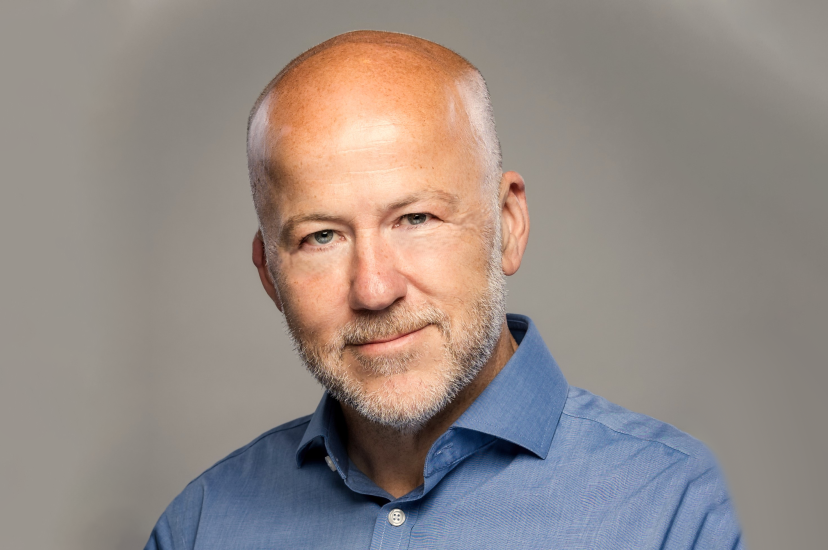 A white, bald man with stubble wearing a blue shirt against a beige background