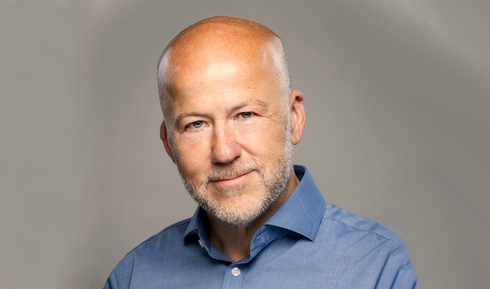 A white, bald man with stubble wearing a blue shirt against a beige background