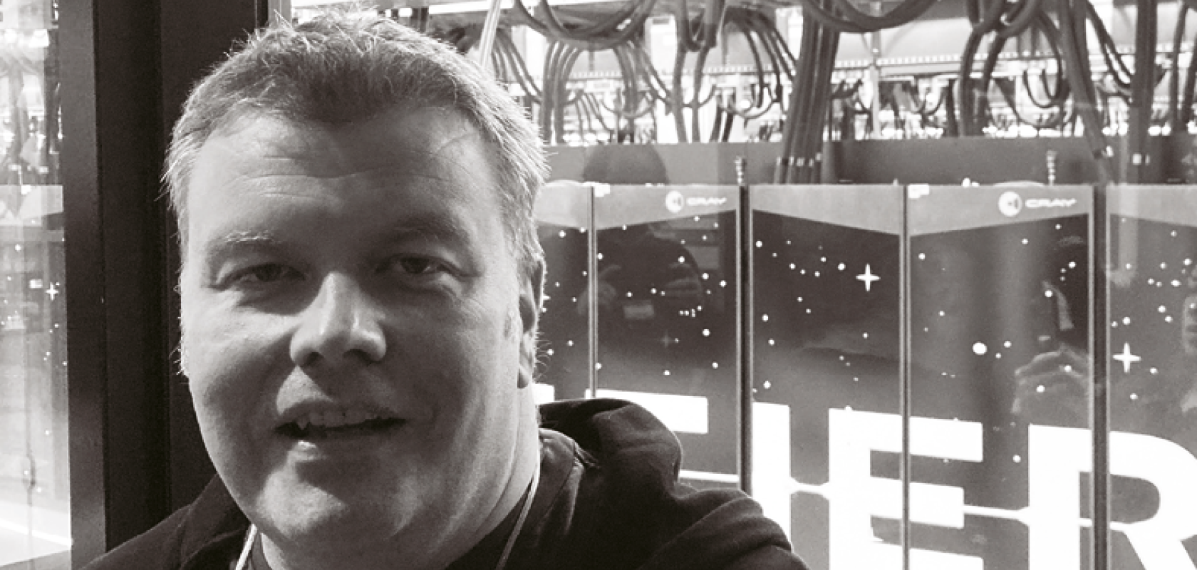 A white man with close-cropped hair stands in front of a window to a server room