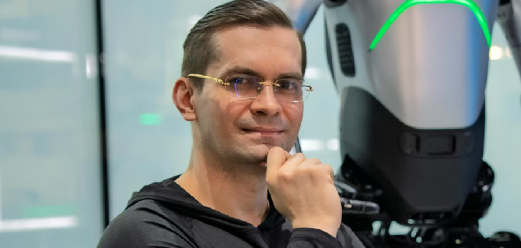 A white man with short dark hair and glasses sits with his finger rested on his chin