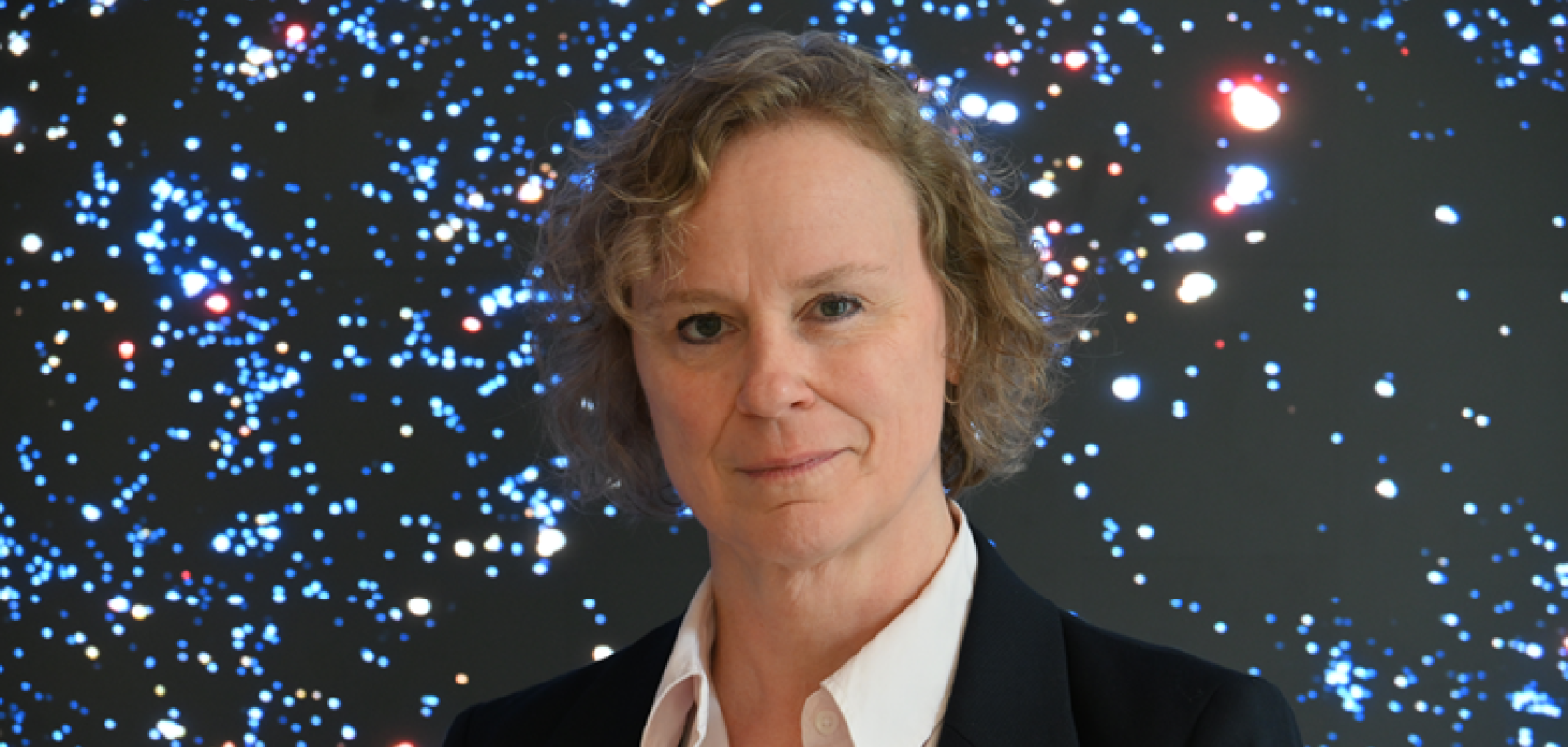 A white woman with blonde, curlyhair and wearing a white shirt and dark jacket, stands in front of a starfield