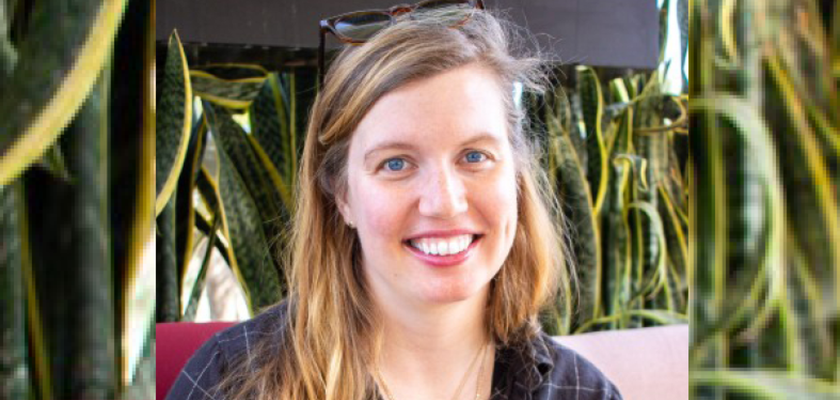 A white woman with fair hair and wearing a blue shirt sits in front of a leafy background