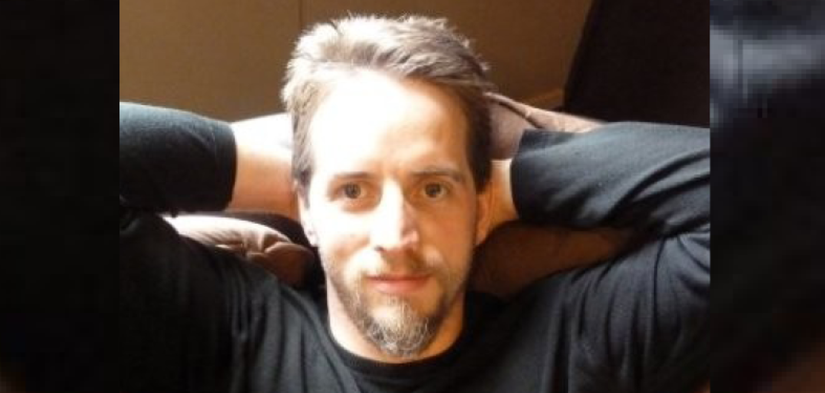 A white  man with fair hair and a beard sits with his arms behind his head