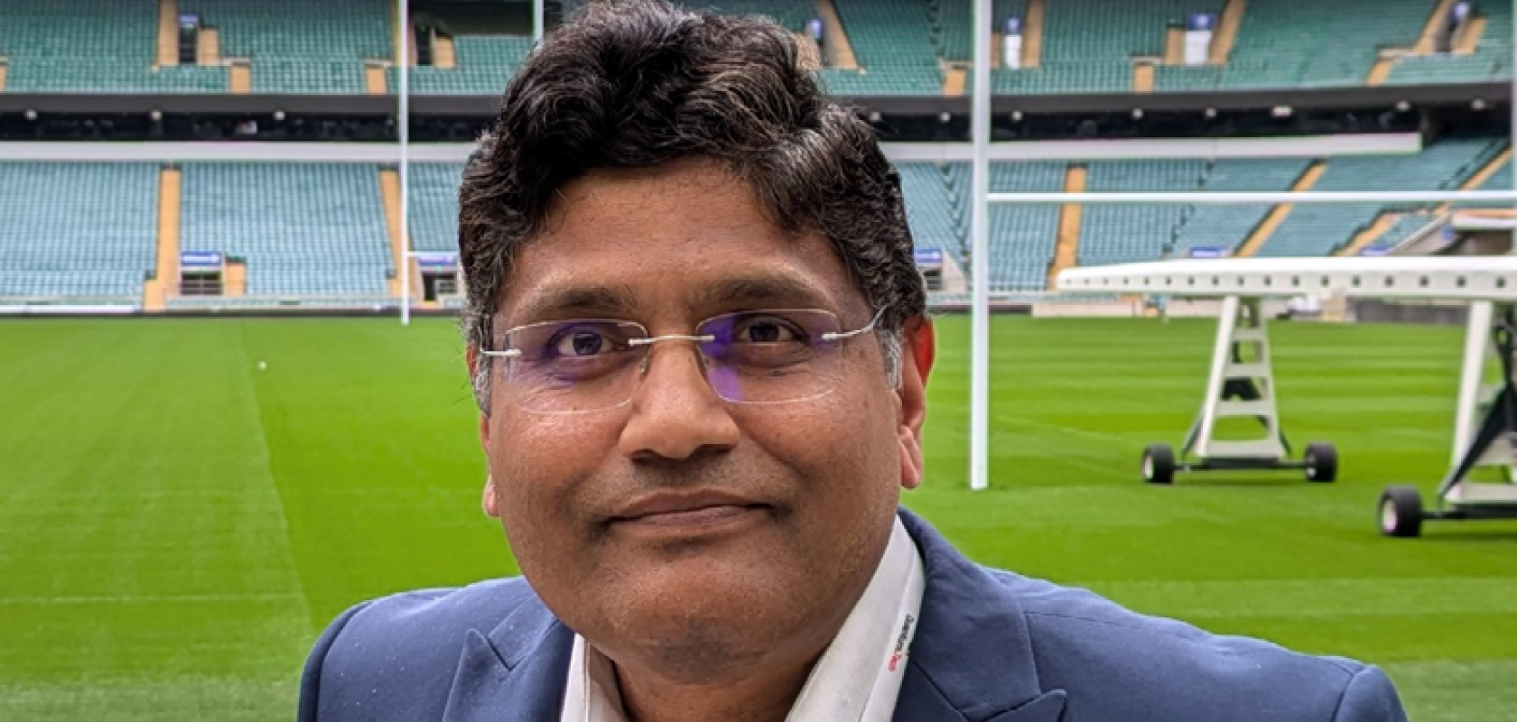 A Asian man with dark hair wearing a suit jacket sands in front of a rugby pitch