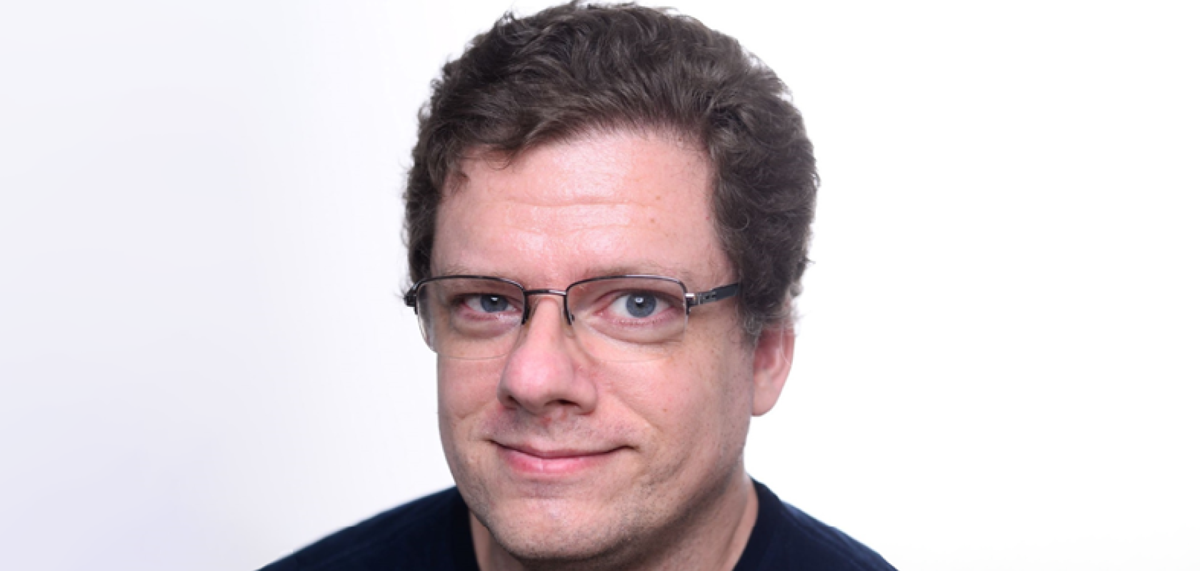 A white man with glasses and curly hair wearing a blue t-shirt
