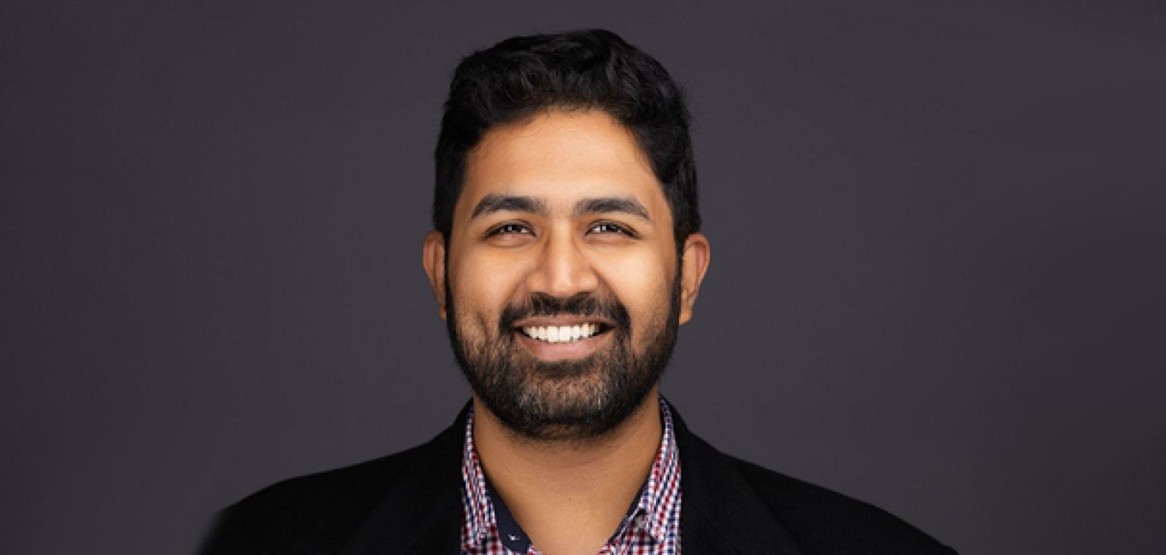 An Asian man with dark hair and beard wearing a dark jacket and ginghamshirt