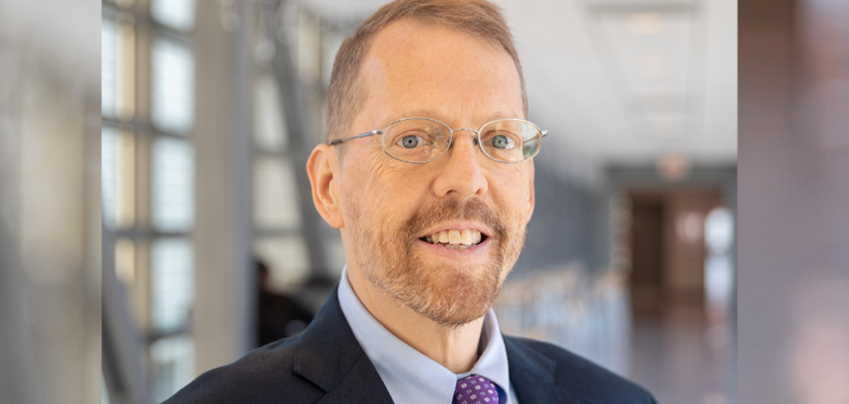 A white man with fair hair and a beard wearing a suit in a brightly-lit corridor