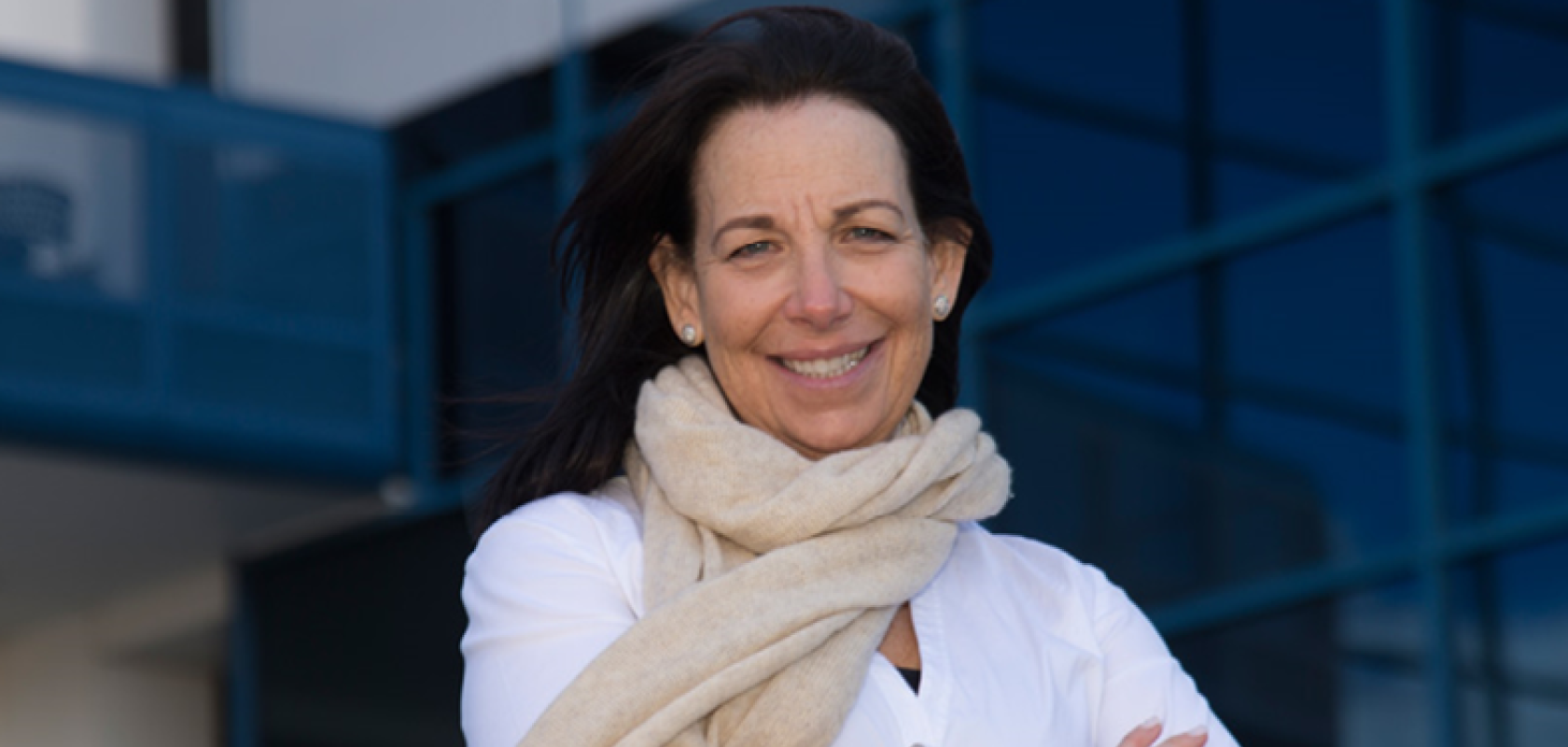 A white woman with brown hair and wearing a white shirt and cream scarf stands in front of a glass building