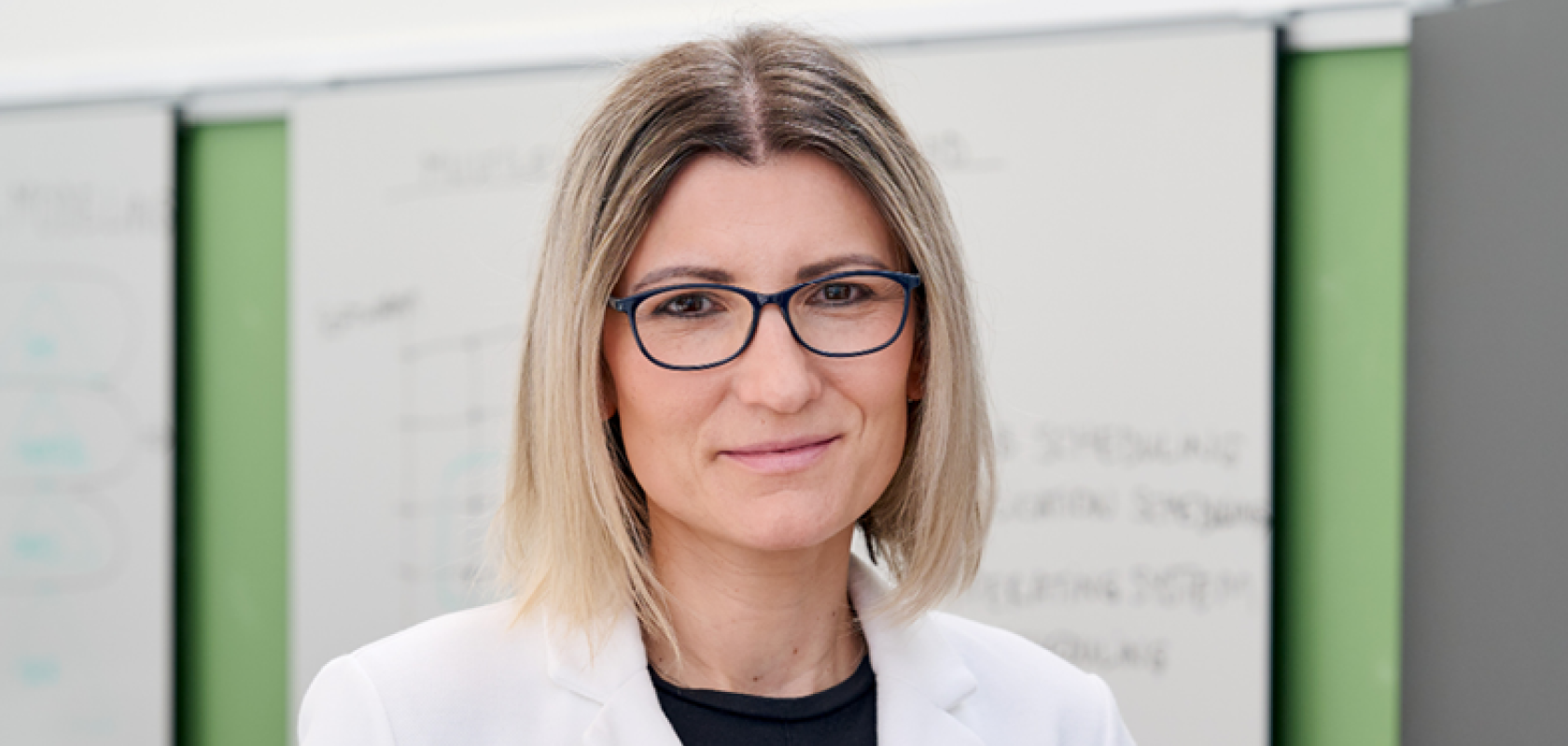 A blonde woman with glasses wearing a lab coat stands in front of a whiteboard