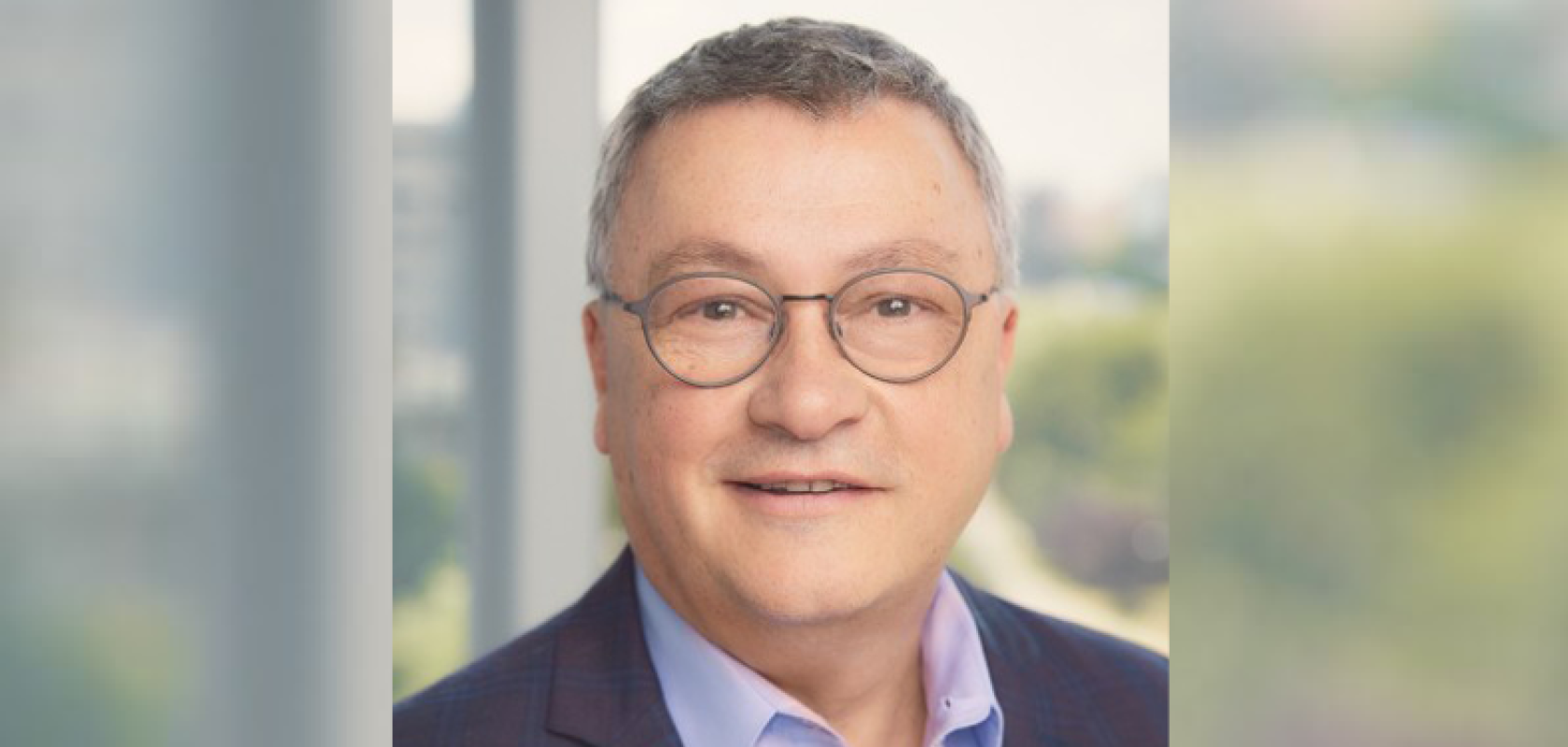 A white man with greying hair and glasses is wearing a suit and standing in front of a window