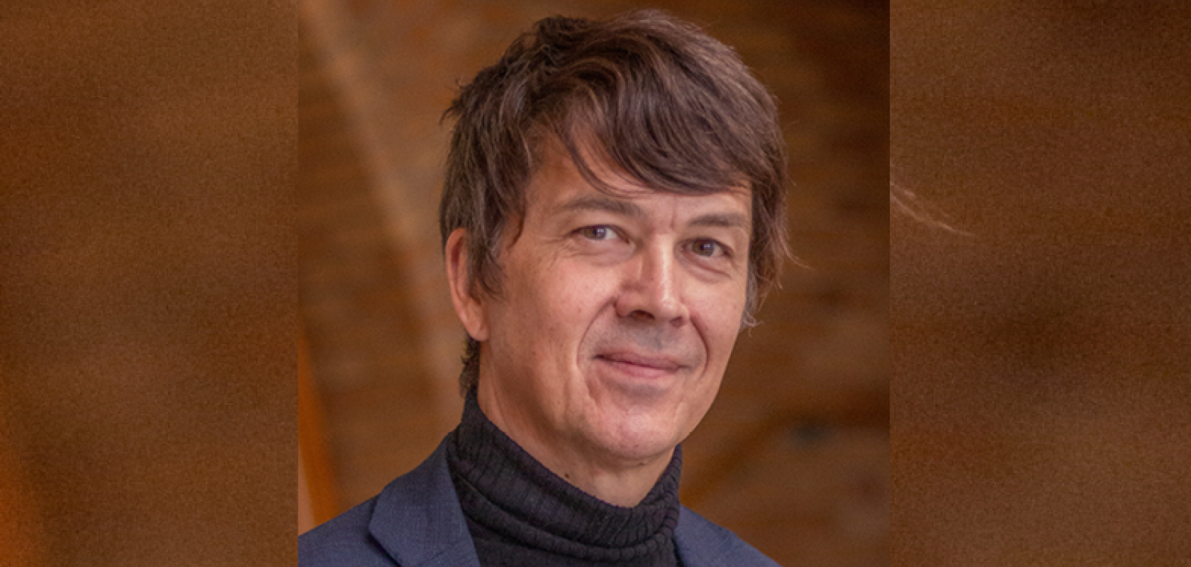 A white man with greting hair and wearing a grey jacket stands in front of a brown background