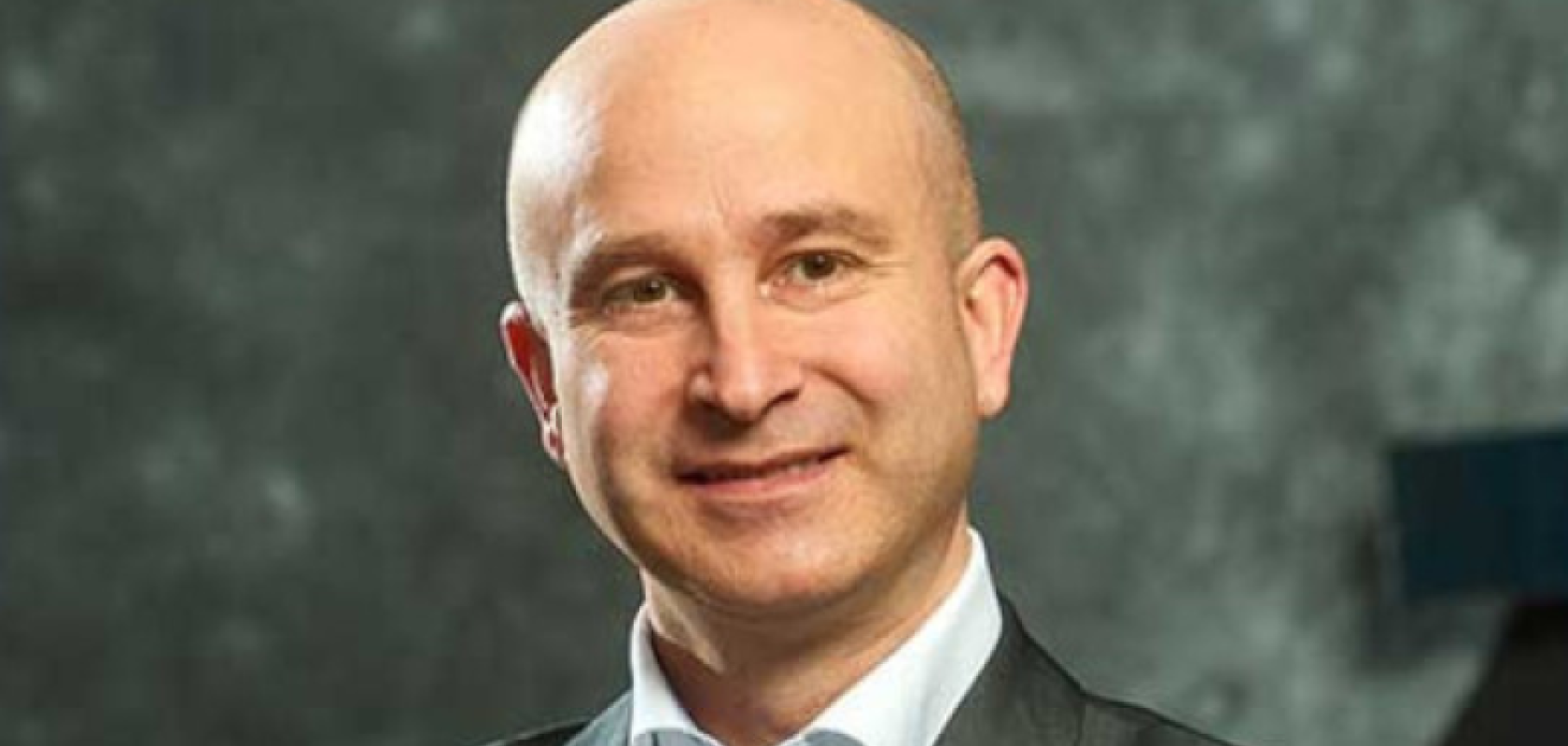 A bald white man wearing a suit stands before a green background 